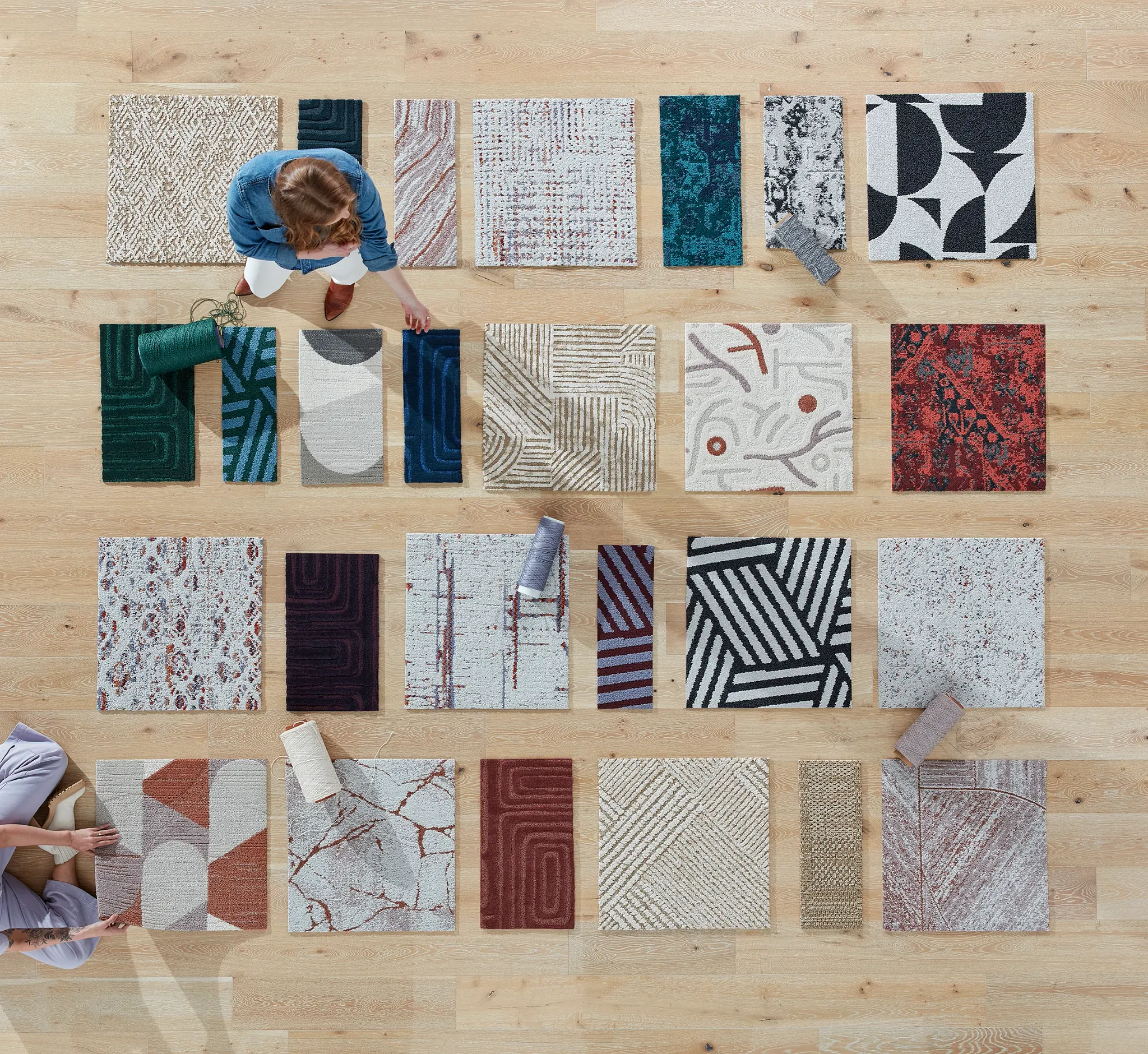 Overhead view of diverse carpet tile swatches in various colors and patterns on a wooden floor with two people arranging them