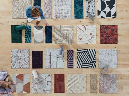 Carpet Tile Swatches Display on Wooden Floor Overhead view of diverse carpet tile swatches in various colors and patterns on a wooden floor with two people arranging them