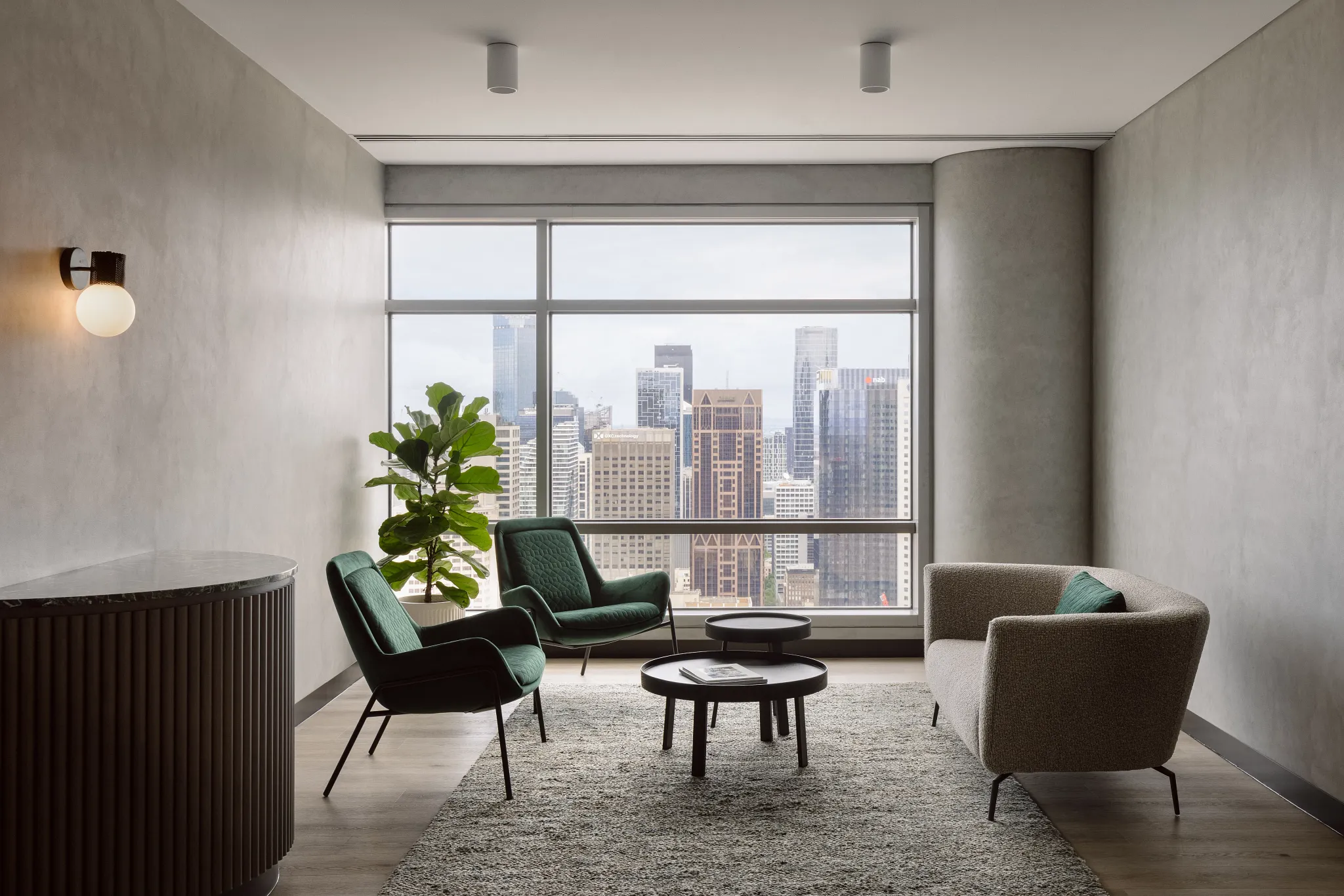 Modern office lounge with city view, featuring green chairs, a gray sofa, and a textured rug on hardwood flooring