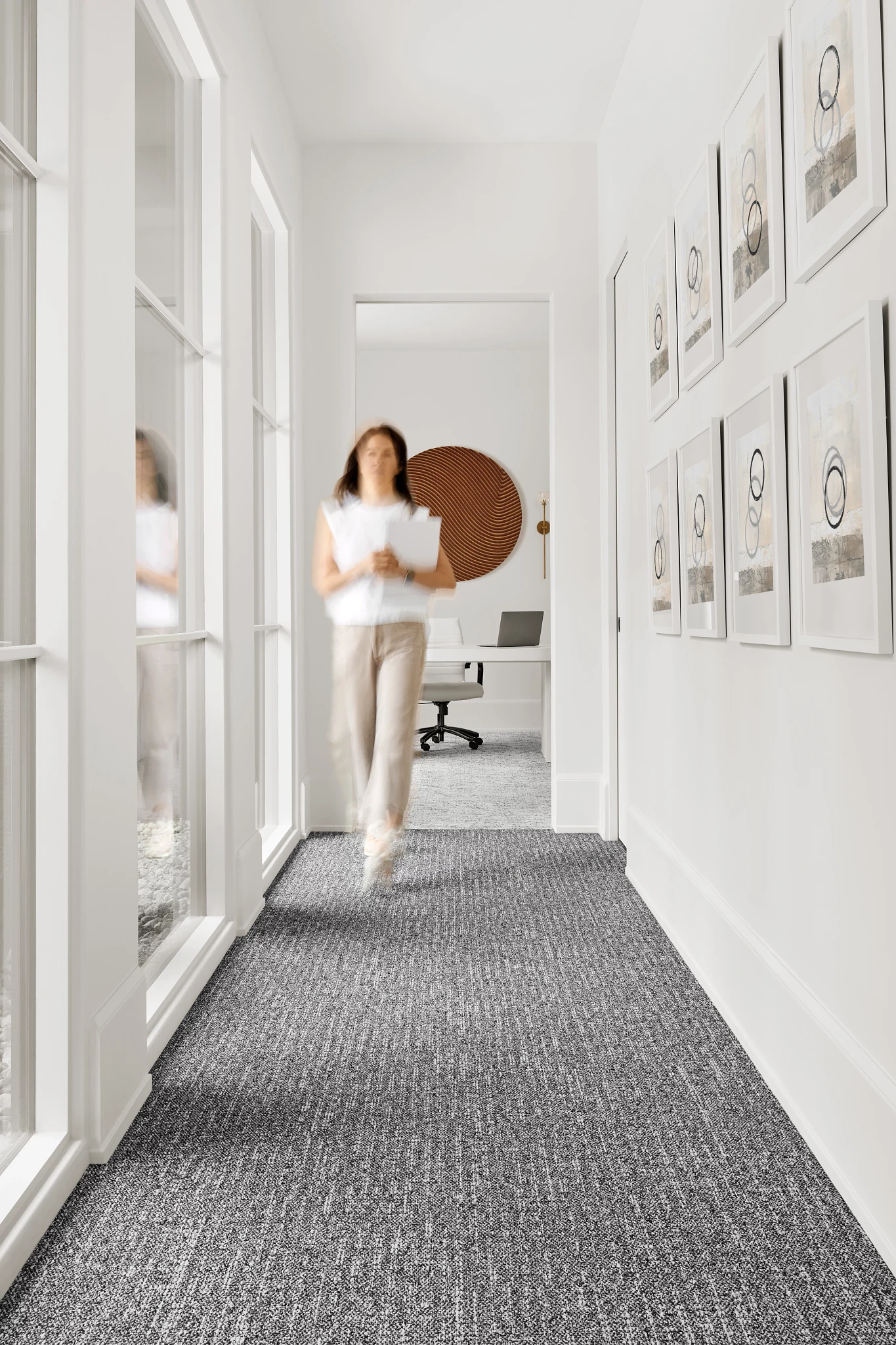 Modern hallway with commercial carpet tile flooring in a neutral gray tone, featuring a woman walking and framed artwork on the walls
