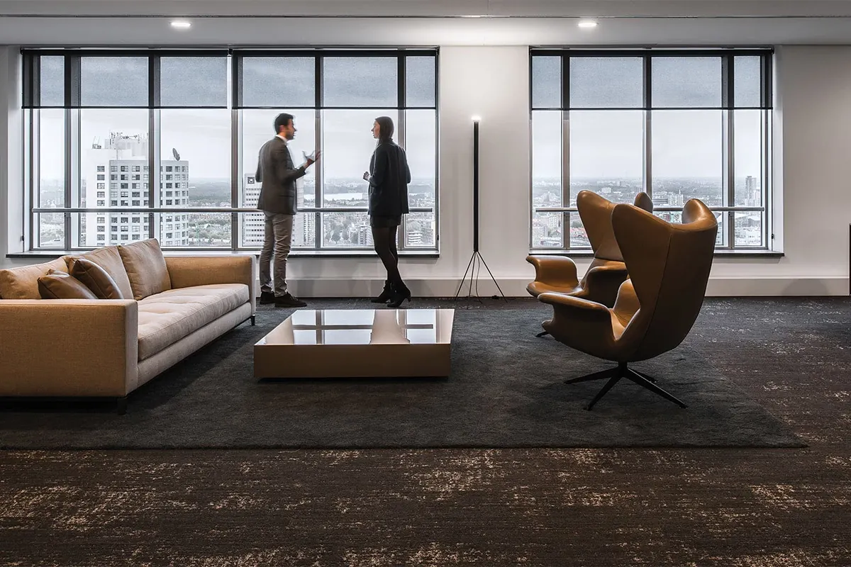 Modern office space with large windows, two people conversing, and dark commercial carpet flooring