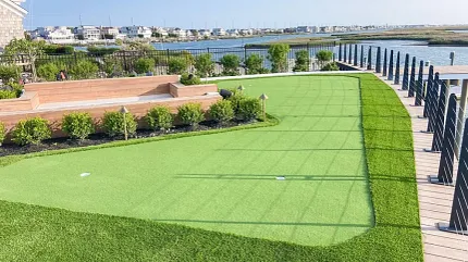 Backyard Putting Green Installation with Waterfront View | Shaw Grass Residential backyard putting green overlooking waterfront homes with elevated wood deck and artificial turf landscaping.