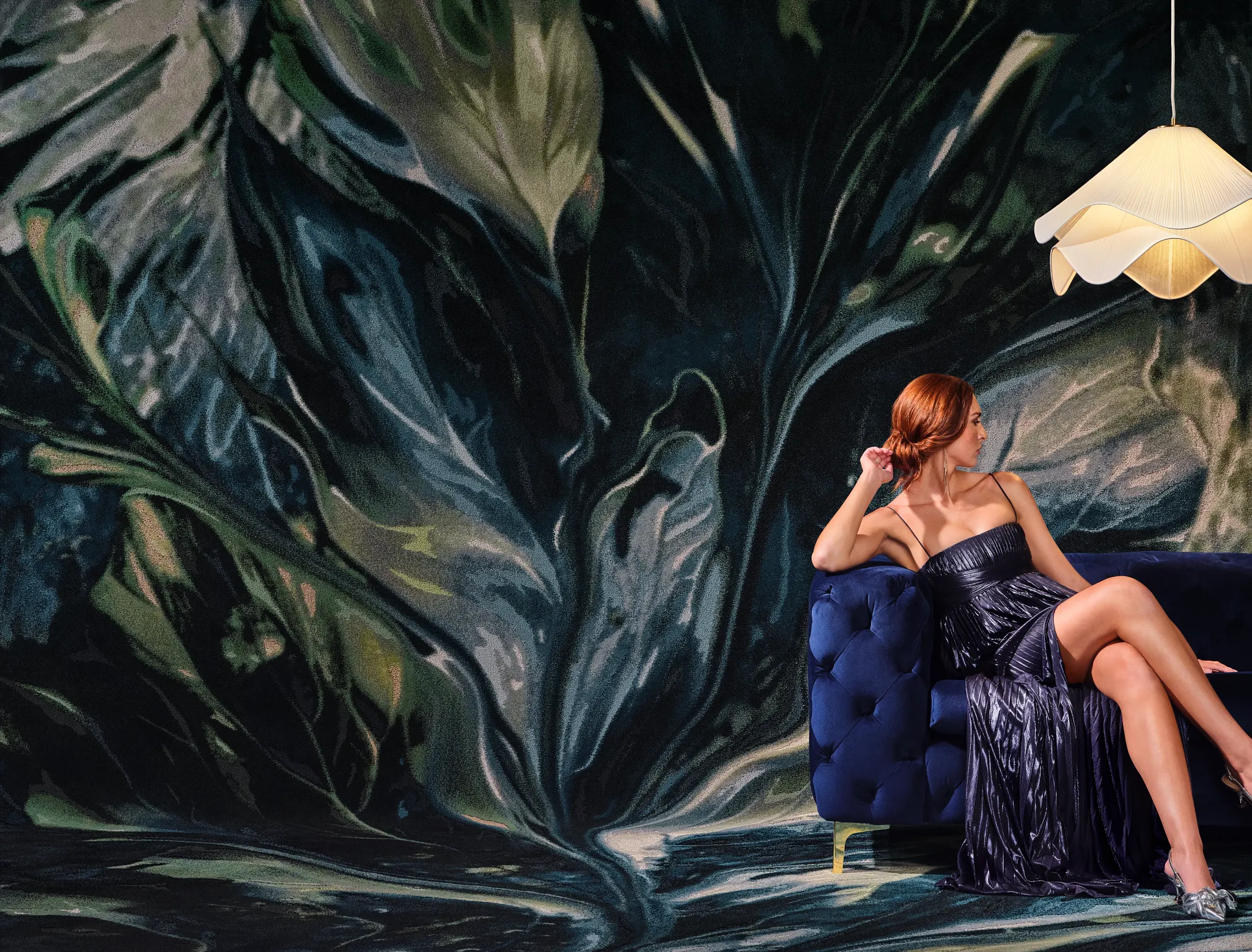 Elegant room scene with a woman in a dark blue dress sitting on a plush blue sofa, featuring a large, artistic leaf-patterned wall mural and a unique pendant light