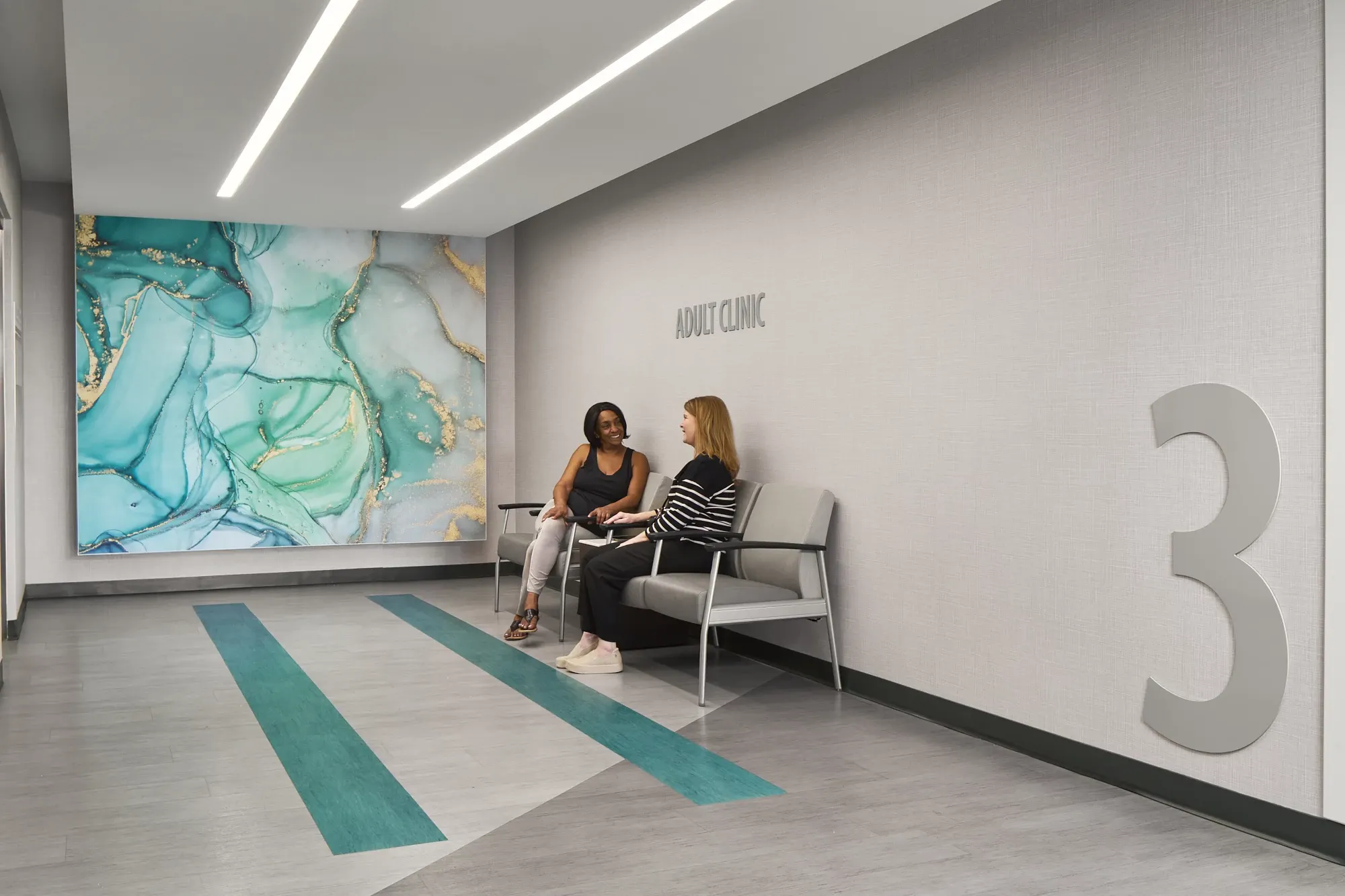 Healthcare facility waiting area with modern design featuring teal and gray flooring, abstract wall art, and two women seated on chairs