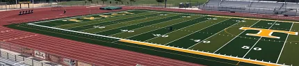 Artificial Turf Football Field with Running Track and Bleachers Football field with artificial turf surrounded by a running track and bleachers under a clear blue sky