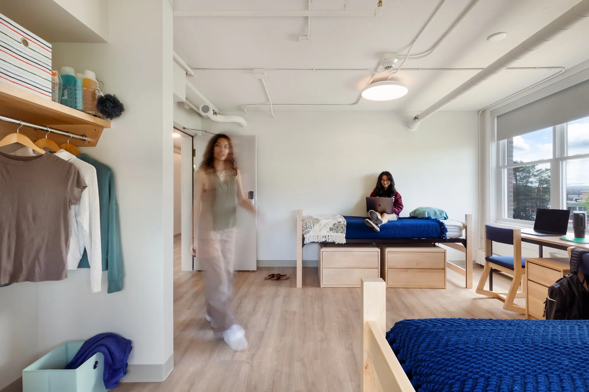 "Higher education dorm room with COREtec flooring, featuring a young woman sitting on a bed with a laptop and another person walking, showcasing 48.03x6.92 inch multilayer SPC with acoustic backing."