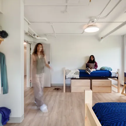 "Higher Education Dorm Room with COREtec Flooring - 48.03x6.92 inch Multilayer SPC" "Higher education dorm room with COREtec flooring, featuring a young woman sitting on a bed with a laptop and another person walking, showcasing 48.03x6.92 inch multilayer SPC with acoustic backing."