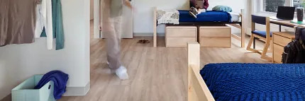"Higher Education Dorm Room with COREtec Flooring - 48.03x6.92 inch Multilayer SPC" "Higher education dorm room with COREtec flooring, featuring a young woman sitting on a bed with a laptop and another person walking, showcasing 48.03x6.92 inch multilayer SPC with acoustic backing."