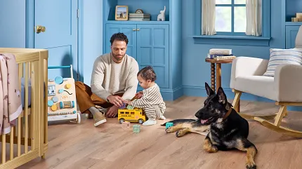 Light Wood Look Luxury Vinyl Flooring in Blue Nursery with Dad, Baby, and Dog A man and a child playing with toys on the floor of a blue-themed room accompanied by a German Shepherd dog