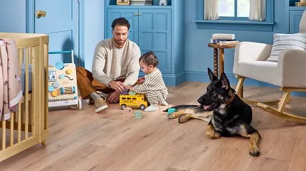Light Wood Look Luxury Vinyl Flooring in Blue Nursery with Dad, Baby, and Dog A man and a child playing with toys on the floor of a blue-themed room, accompanied by a German Shepherd dog. The room includes a crib, a toy walker, shelves with books and decor, a window with light coming through, and luxury vinyl plank flooring
