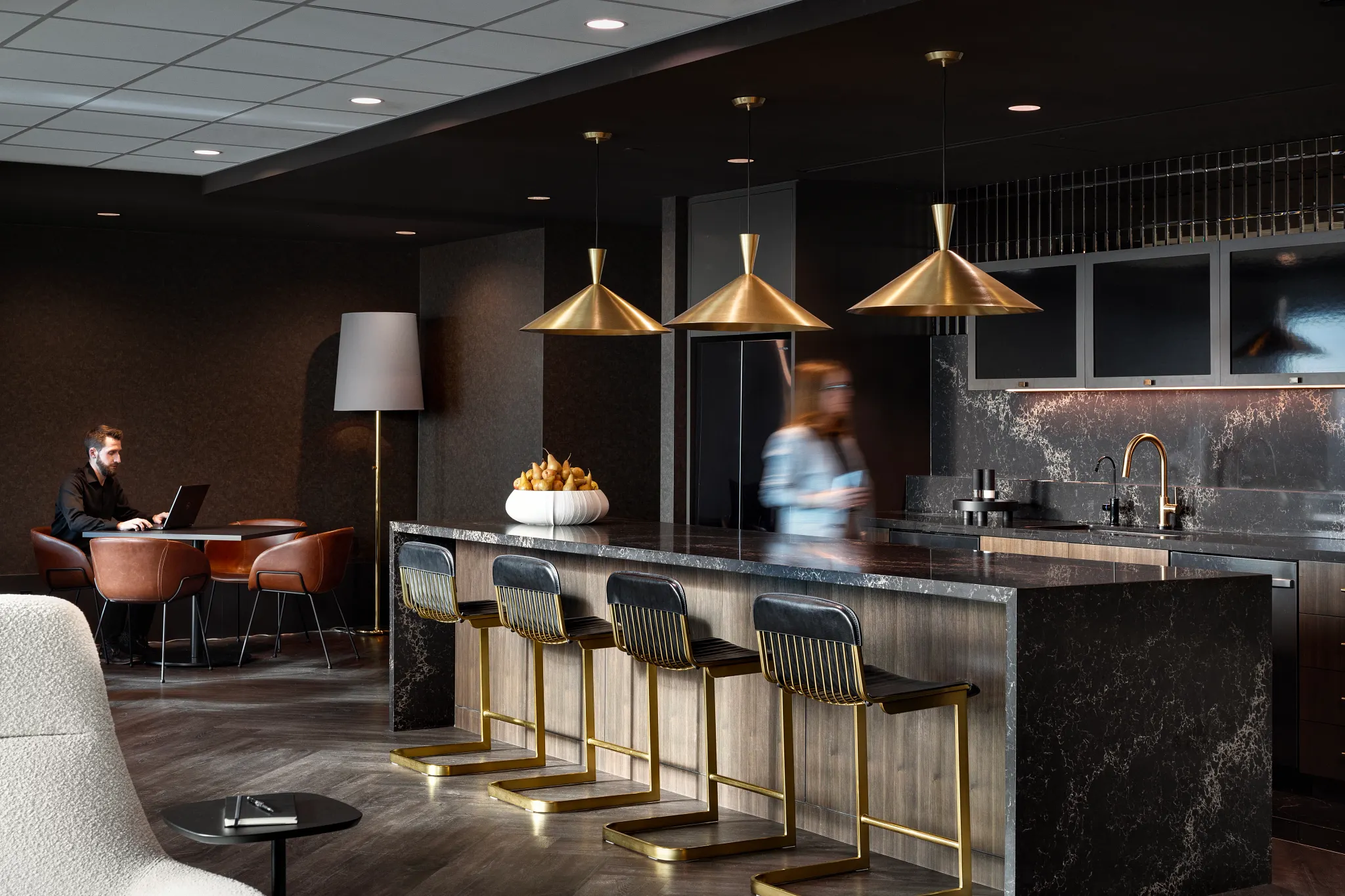 Modern workplace lounge with sleek bar area, gold pendant lights, and stylish seating on dark wood flooring