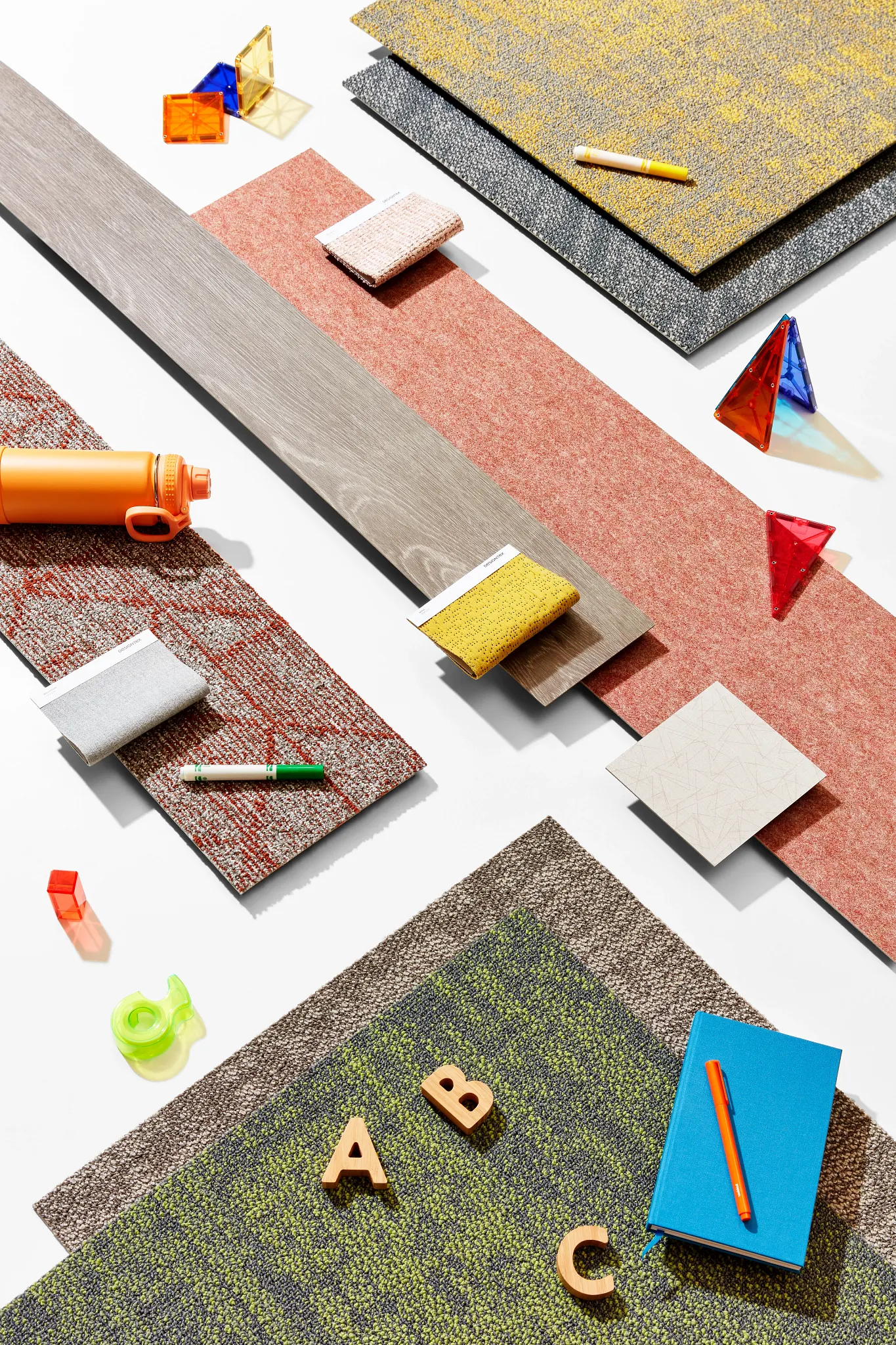 Assorted flooring swatches including commercial carpet tiles in yellow, gray, red, and green, with educational items like a notebook, pen, and geometric shapes
