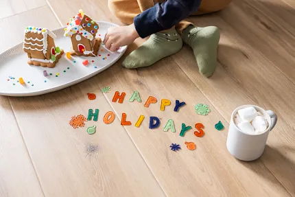 Light Wood Look Luxury Vinyl Plank Flooring - Holiday Scene Child decorating gingerbread houses on light wood look luxury vinyl plank flooring with "Happy Holidays" letters and a mug of hot chocolate