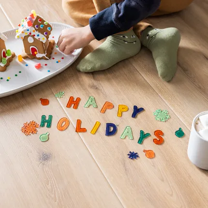 Light Wood Look Luxury Vinyl Plank Flooring - Holiday Scene Child decorating gingerbread houses on light wood look luxury vinyl plank flooring with "Happy Holidays" letters and a mug of hot chocolate