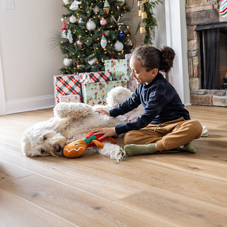 Girl on floor playing with a white dog with a dog toy in front of a Christmas tree with presents under it and a fireplace with stockings hanging