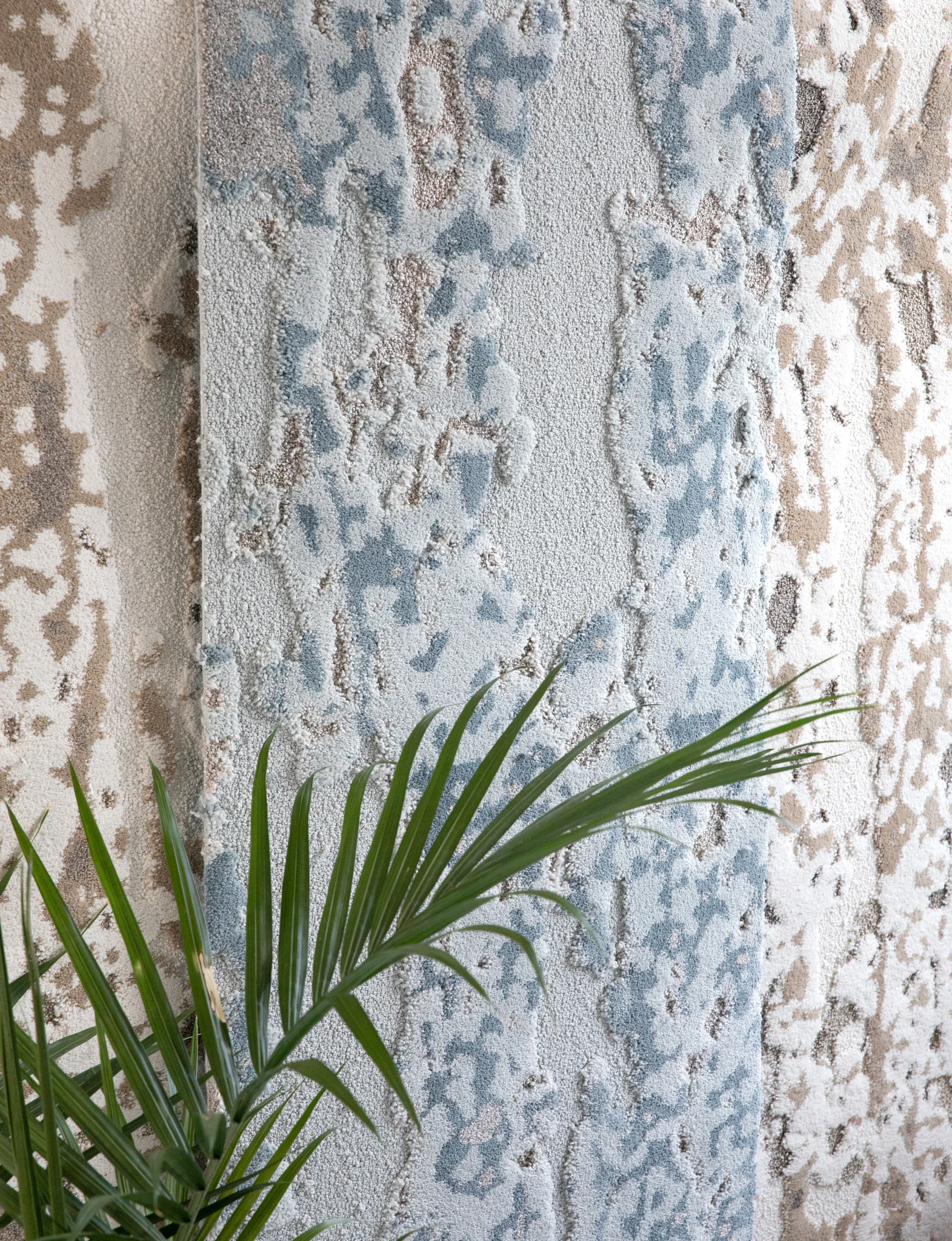 Textured carpet swatch in blue, beige, and cream colors with a leafy plant in the foreground