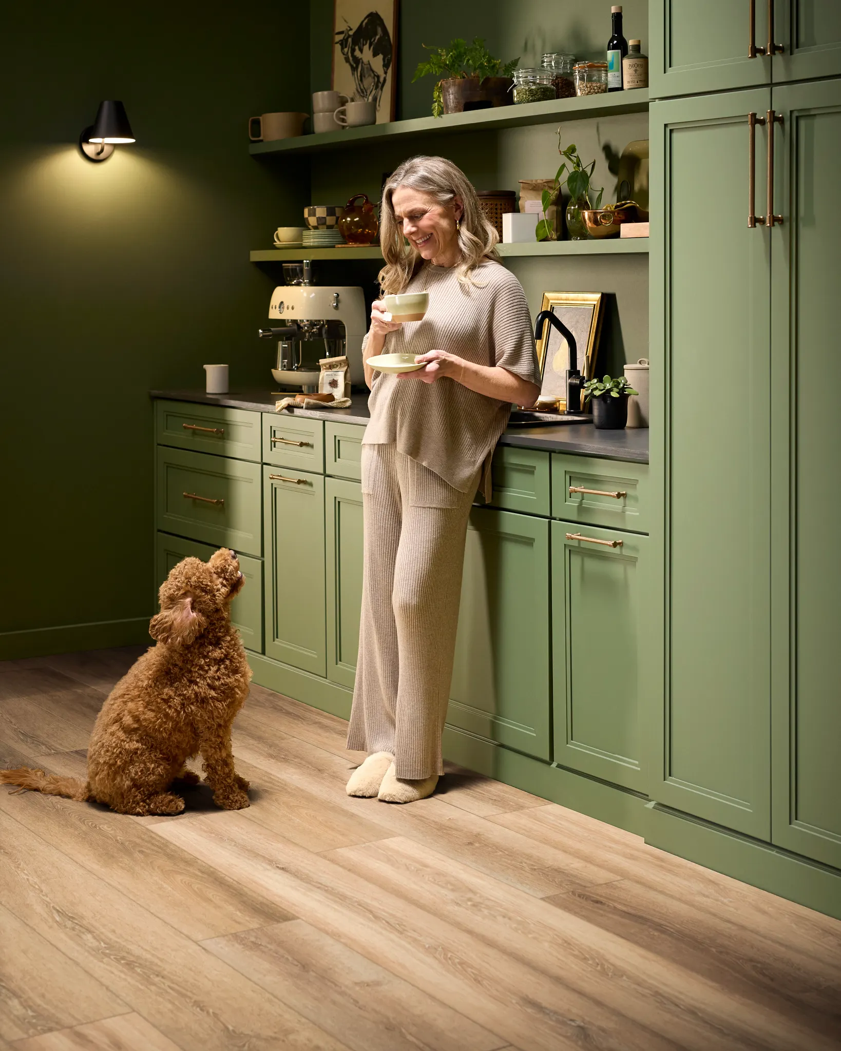 Woman in cozy kitchen with green cabinets and luxury vinyl flooring, holding a cup while a dog looks up at her