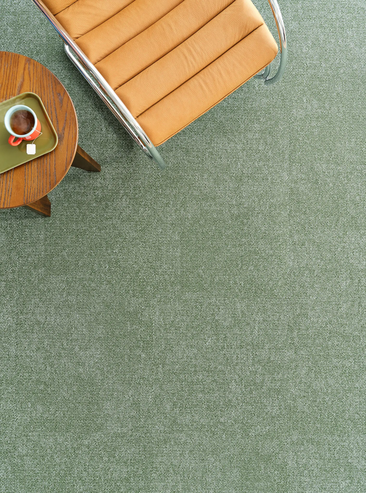 Green Spice Route carpet flooring with subtle tonal variation, paired with a tan leather chair and round timber side table in a contemporary interior setting.