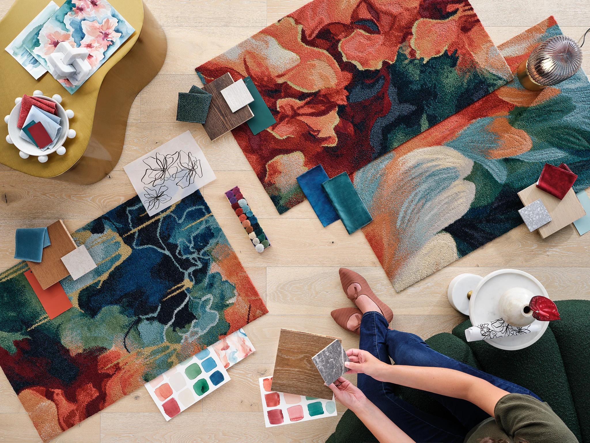 Overhead view of a creative workspace with colorful abstract carpet swatches, paint palettes, and a person holding flooring samples on a light wood floor