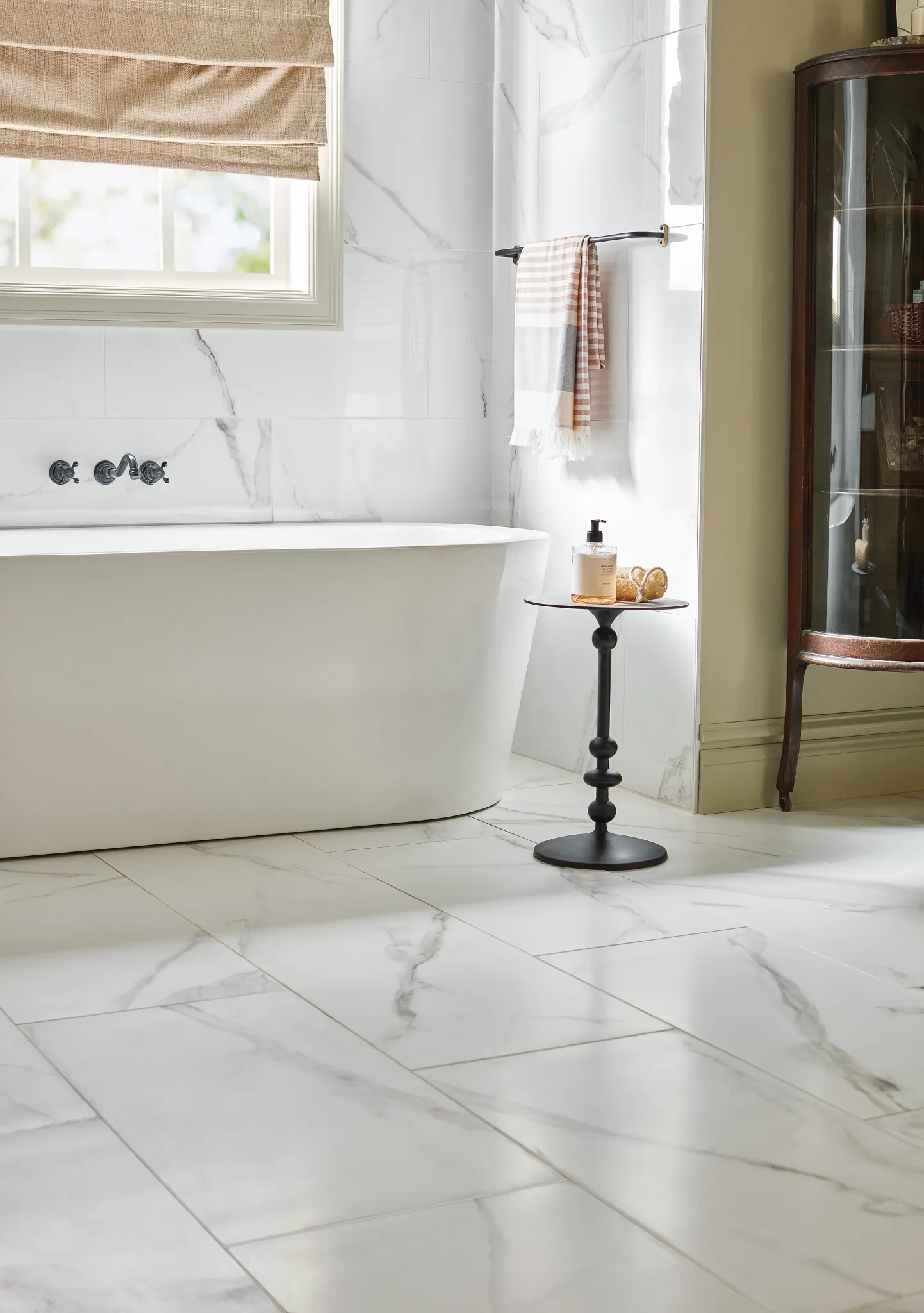 Elegant bathroom with white marble-look tile flooring and walls, featuring a freestanding bathtub and a black side table