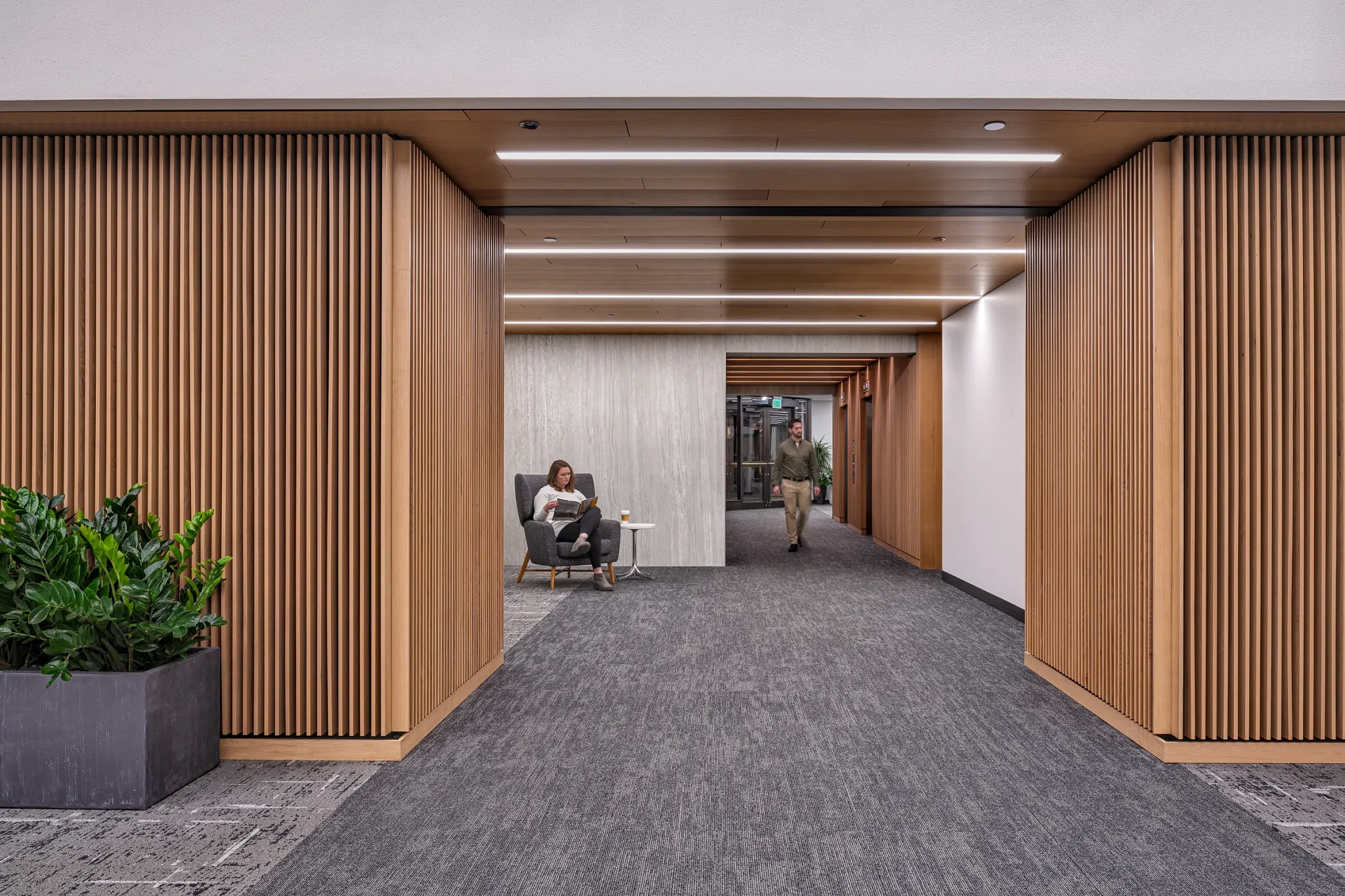 Modern workplace hallway with commercial carpet tile flooring, featuring a woman seated and a man walking, surrounded by wooden paneling and indoor plants