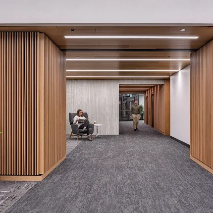 Contemporary Workplace Hallway with Commercial Carpet Tile Flooring Modern workplace hallway with commercial carpet tile flooring, featuring a woman seated and a man walking, surrounded by wooden paneling and indoor plants