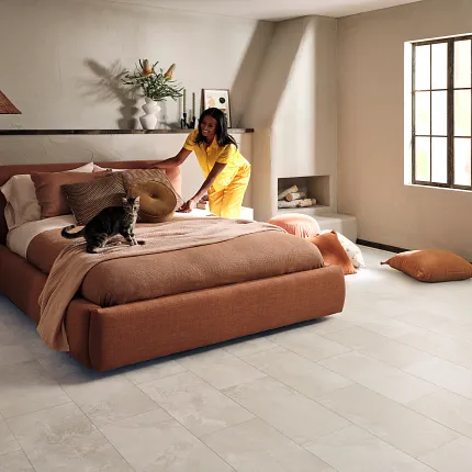 Coretec Mineral Core Residential LVT Flooring in Light Stone Look - 23.62x11.81 inch Modern bedroom with Coretec Mineral Core Residential luxury vinyl tile flooring in a light stone look, featuring a young woman making the bed and a cat sitting on it