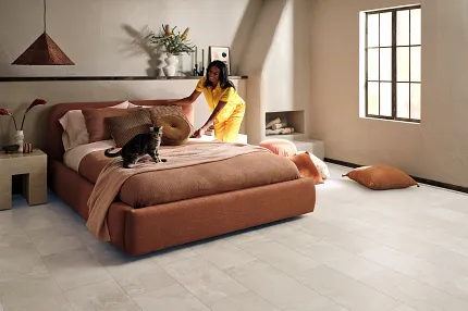 Coretec Mineral Core Residential LVT Flooring in Light Stone Look - 23.62x11.81 inch Modern bedroom with Coretec Mineral Core Residential luxury vinyl tile flooring in a light stone look, featuring a young woman making the bed and a cat sitting on it