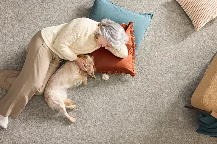Comfortable and durable carpet flooring for everyday home comfort. Cozy living room scene with a woman and dog relaxing on soft, durable carpet flooring, surrounded by colorful pillows in blue, orange, and beige.