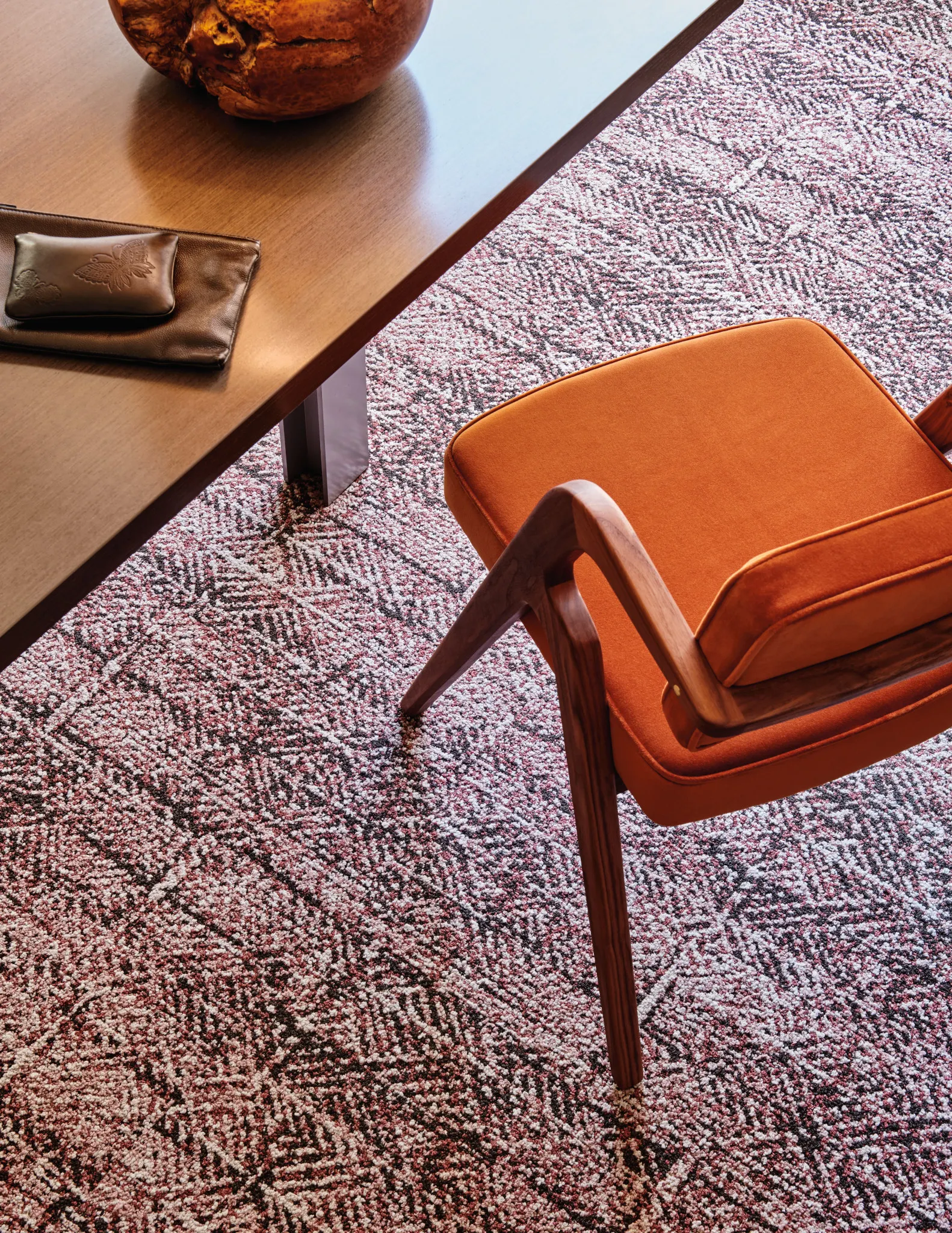 Modern office setting with a wooden table, leather accessories, and an orange chair on a patterned commercial carpet tile in shades of red, black, and
