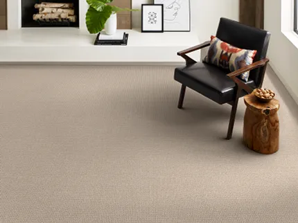 Beige Carpet Flooring in Modern Living Room - 12 ft Width Modern living room with beige carpet flooring, featuring a black leather chair with a colorful pillow and a wooden side table
