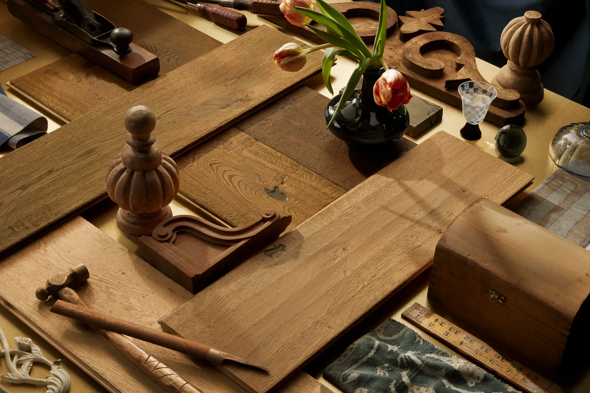 Arrangement of hardwood flooring samples, woodworking tools, carved wood accents, textiles, and decorative objects showcasing craftsmanship, natural materials, and emerging residential design trends.