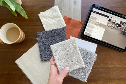 Carpet and Flooring Swatches with Tablet Display on Wooden Table Hand holding carpet swatches in beige, gray, and cream with a tablet displaying flooring options on a wooden table