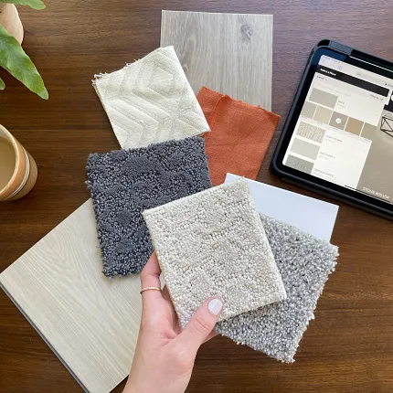 Carpet and Flooring Swatches with Tablet Display on Wooden Table Hand holding carpet swatches in beige, gray, and cream with a tablet displaying flooring options on a wooden table