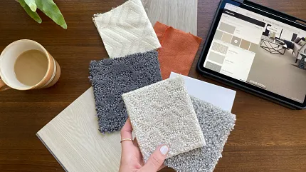 Carpet and Flooring Swatches with Tablet Display on Wooden Table Hand holding carpet swatches in beige, gray, and cream with a tablet displaying flooring options on a wooden table