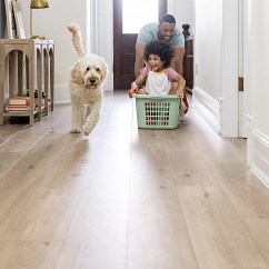 Man pushing child across COREtec vinyl flooring in a laundry basket with cream colored dog, people laughing