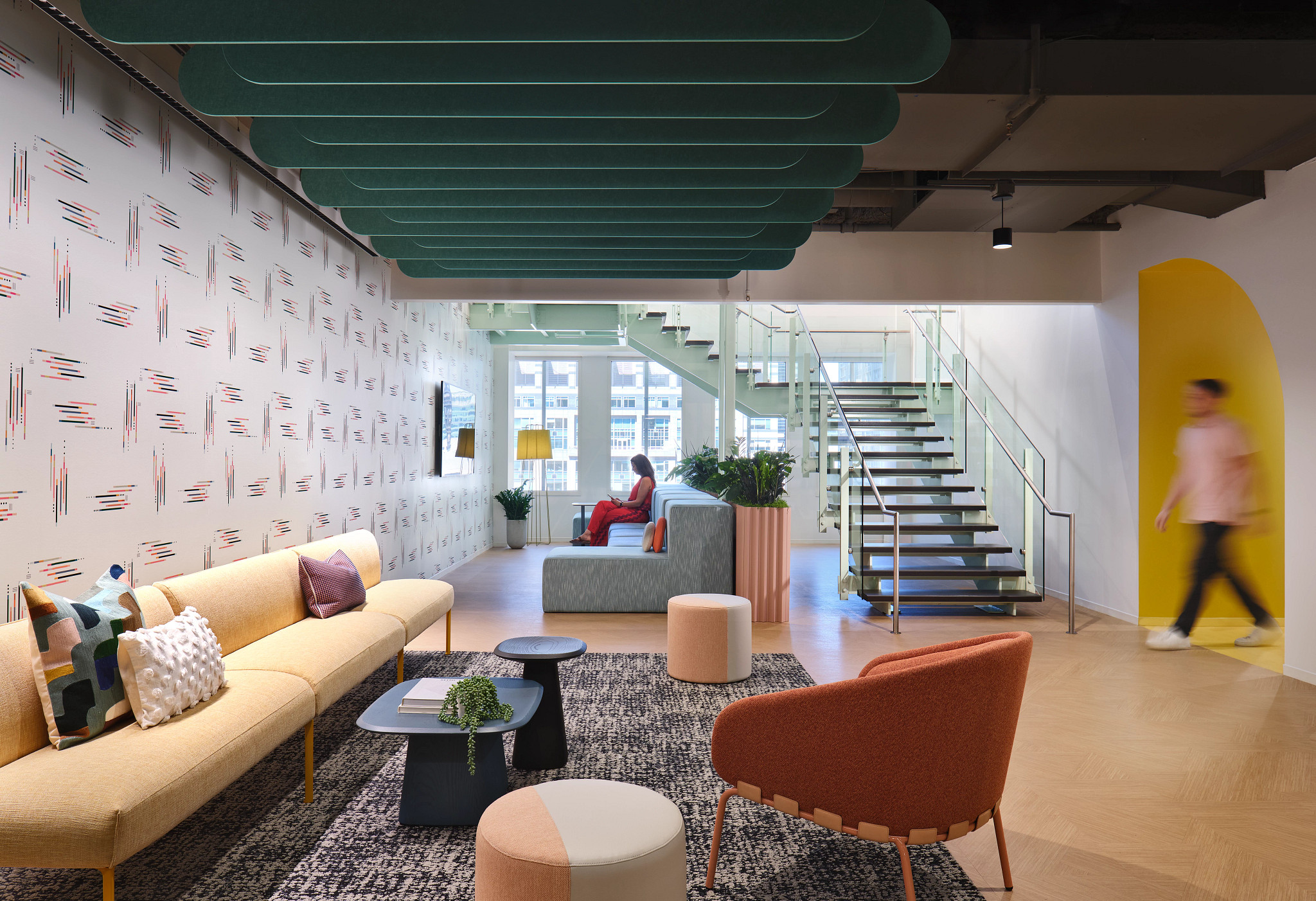 Modern workplace lounge with heavy commercial luxury vinyl tile flooring, featuring 18x18 inch tiles, contemporary furniture, and a person walking by a staircase