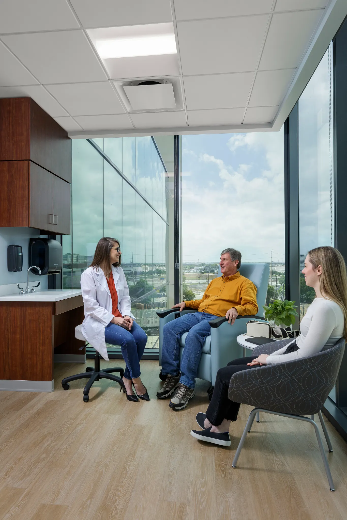 Healthcare facility room with light wood flooring, featuring a healthcare professional and two individuals seated in a consultation setting