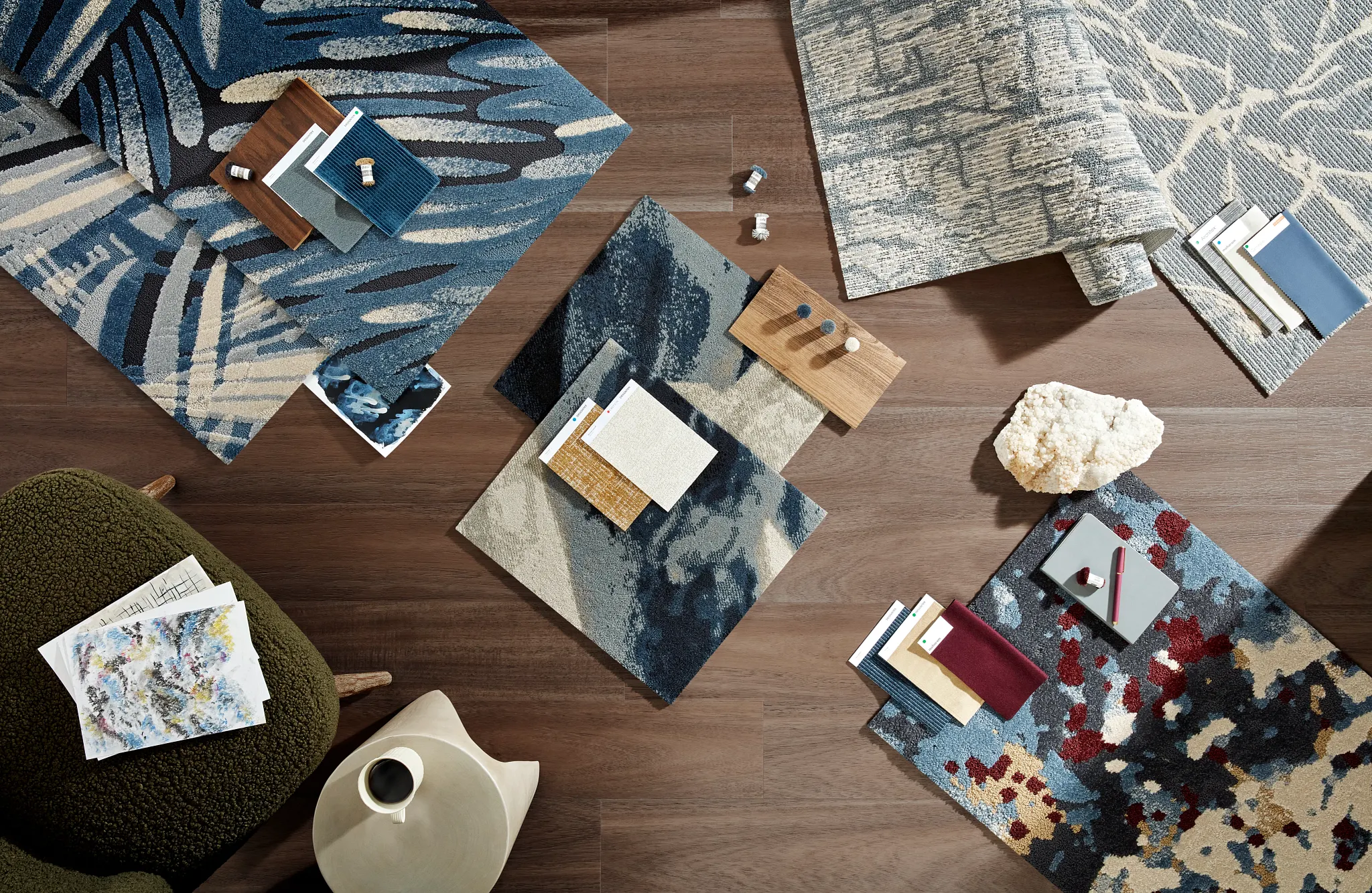Vista superior de muestras de alfombra comercial en tonos azul, beige y rojo sobre un piso de madera, acompañadas de materiales de diseño y una silla verde.