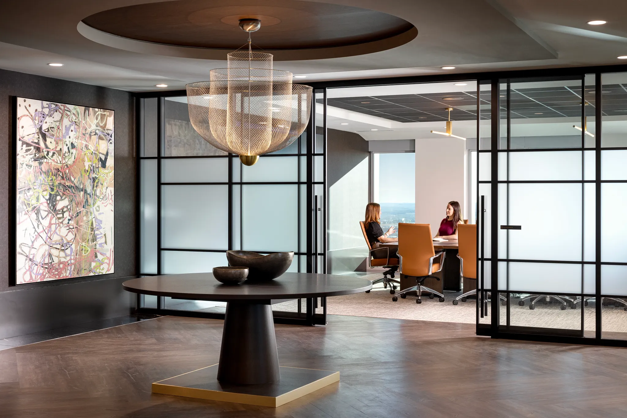 Modern workplace interior with elegant chandelier and abstract art, featuring dark wood flooring and glass partitioned meeting room with two women in discussion