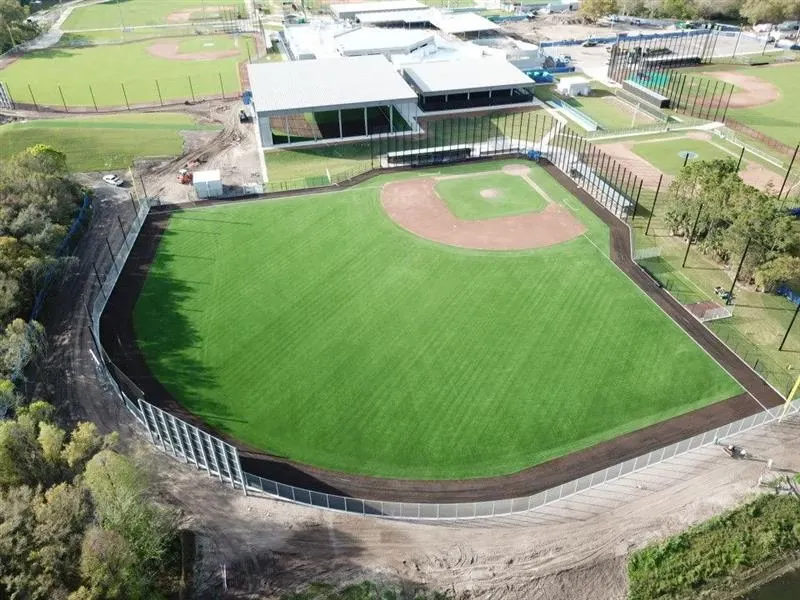 Aerial view of a baseball field with artificial turf surrounded by fencing and adjacent buildings