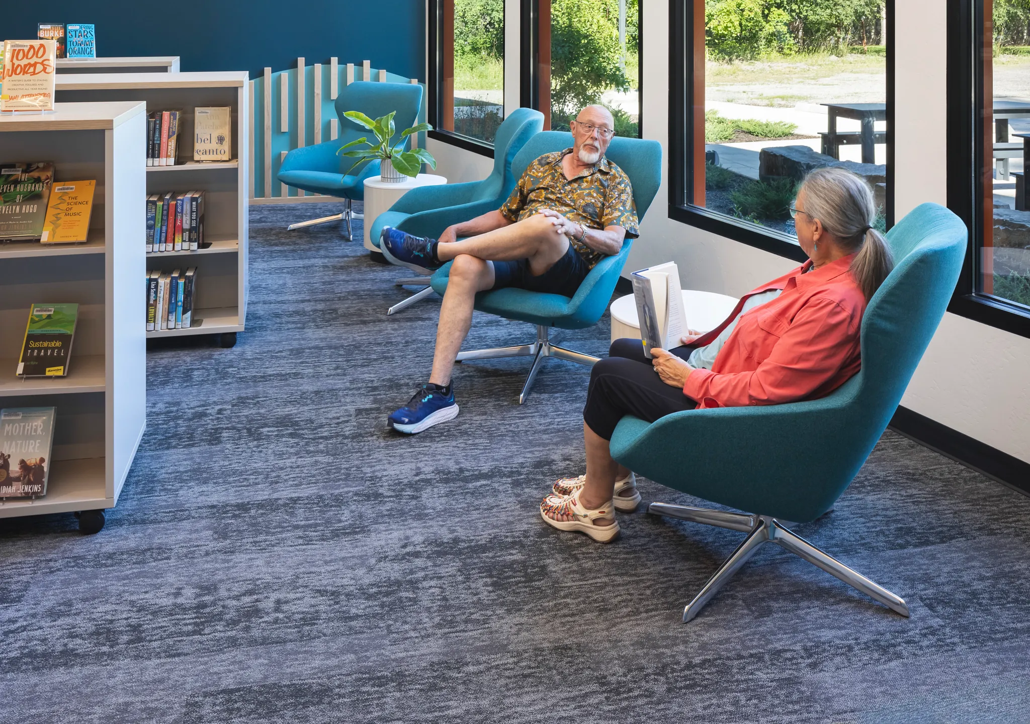 Education facility with commercial carpet tile flooring in a library setting, featuring blue and gray tones, 9x36 inch dimensions, with people reading and relaxing in teal chairs near large windows.