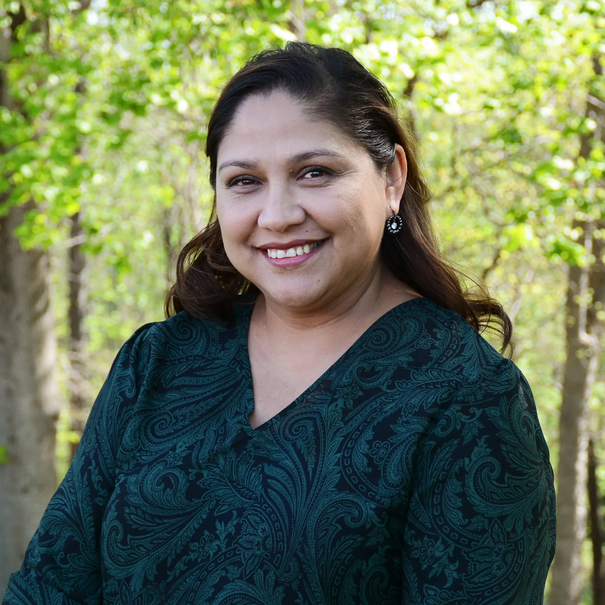 A business style headshot of a woman with a dark blue-green dress and brown hair with trees in the background