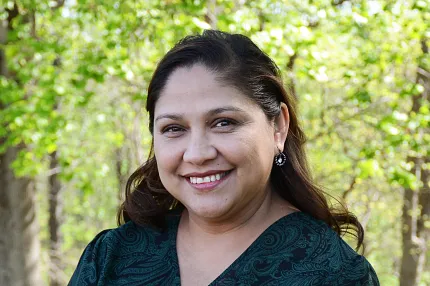 A business style headshot of a woman with a dark blue-green dress and brown hair with trees in the background