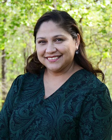 A business style headshot of a woman with a dark blue-green dress and brown hair with trees in the background