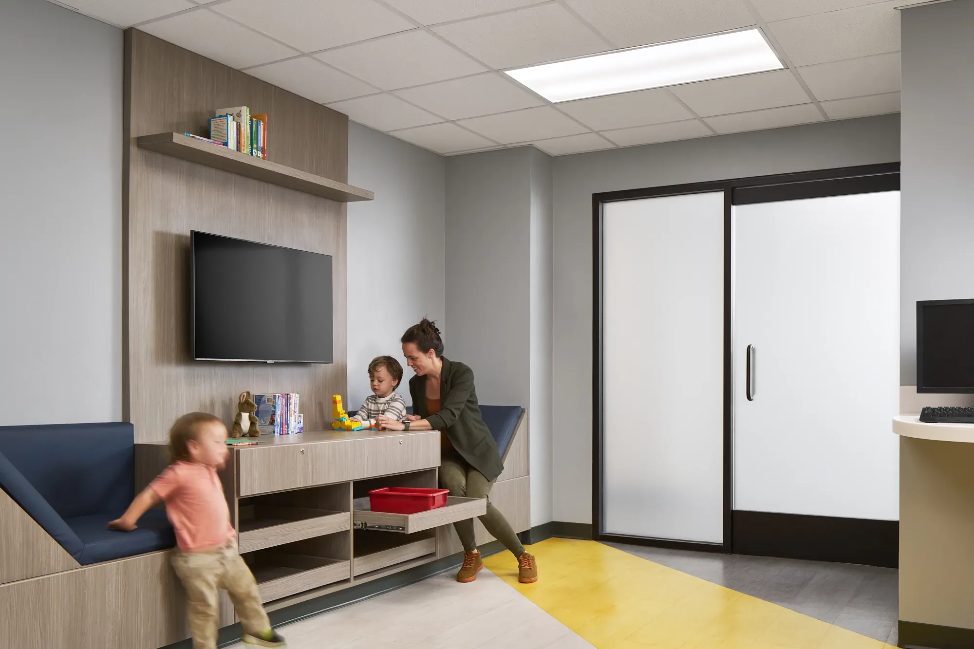 Modern healthcare facility waiting area with yellow and gray flooring, featuring a woman and children interacting near a TV and storage unit