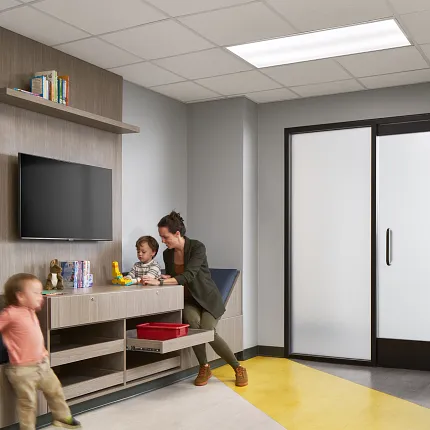 Healthcare Facility Waiting Area with Yellow and Gray Flooring Modern healthcare facility waiting area with yellow and gray flooring, featuring a woman and children interacting near a TV and storage unit