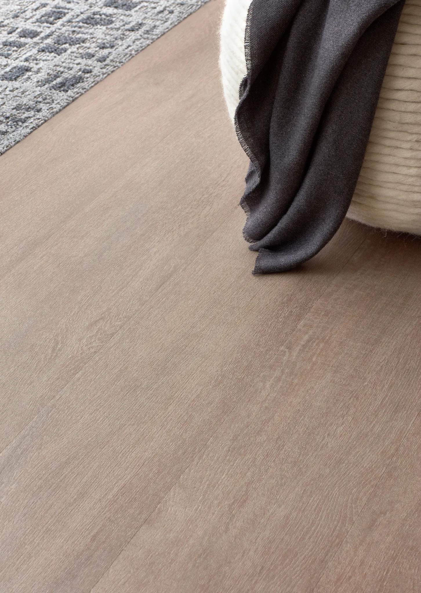 Close-up of light brown hardwood flooring with a textured surface, featuring a dark gray fabric draped over a cream-colored ottoman.