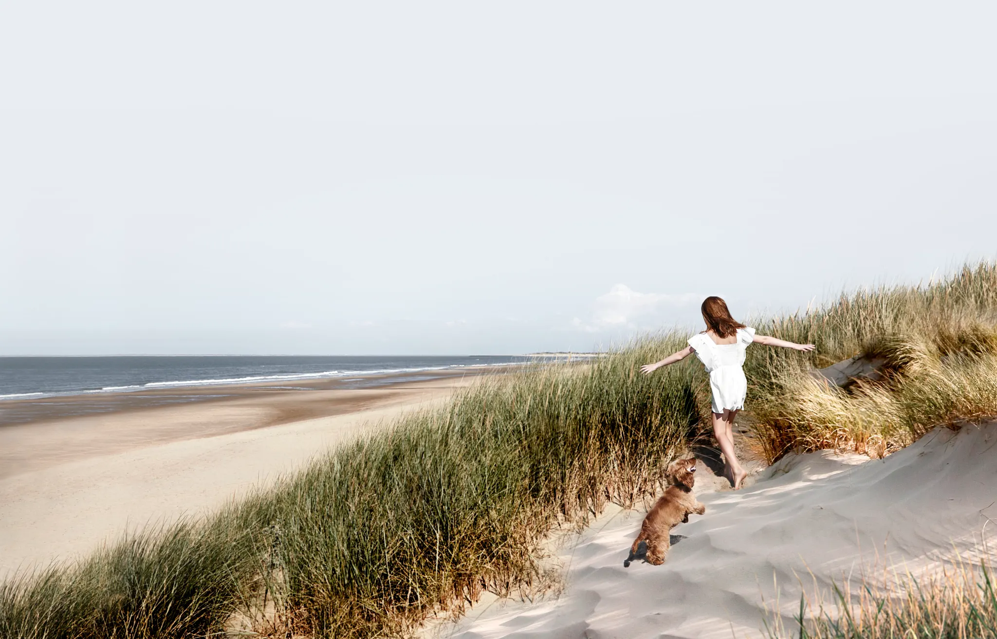 Woman and dog walking on a sandy beach with grassy dunes