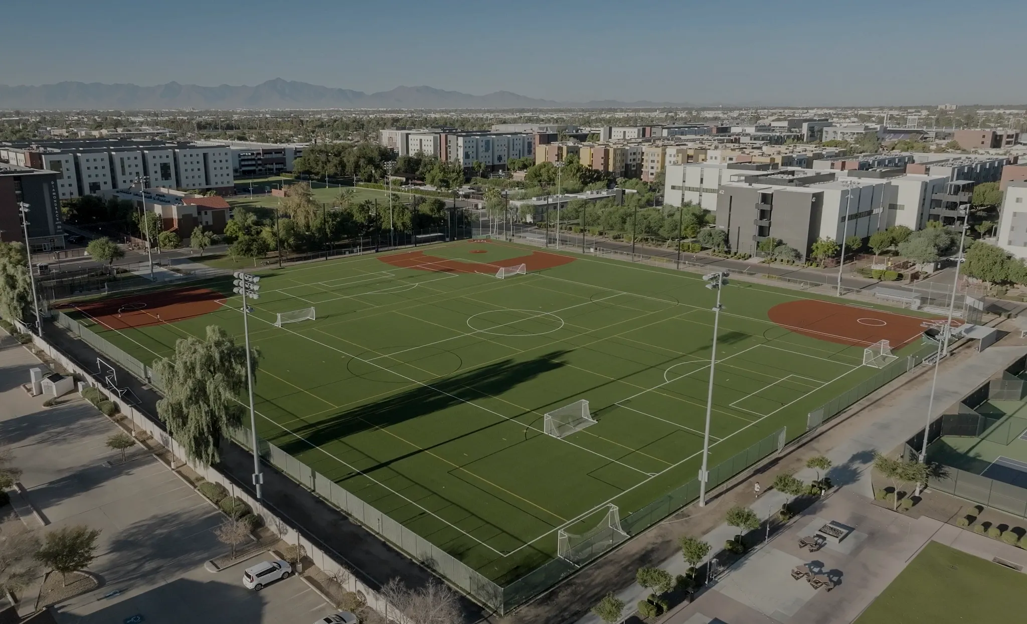 Aerial view of a sports complex featuring multiple artificial turf soccer fields surrounded by urban buildings and mountains in the background